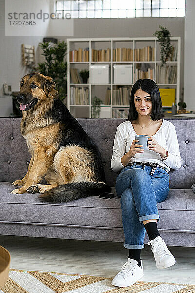Junge Frau hält Kaffeetasse  während sie mit Hund auf dem Sofa zu Hause sitzt