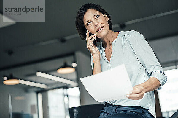 Unternehmerin mit Dokument  die im Büro mit ihrem Smartphone telefoniert