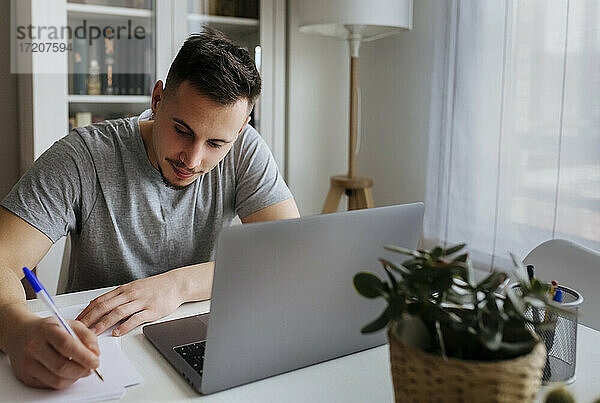 Männlicher Unternehmer  der mit einem Laptop im Büro zu Hause sitzt und auf Papier schreibt