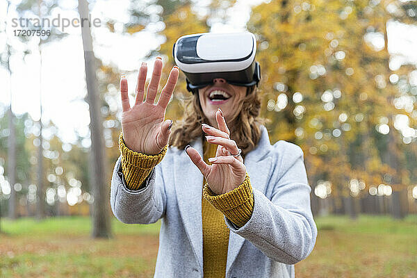 Lächelnde Frau mit Virtual-Reality-Headset  die ihre Hände ausstreckt  während sie im Wald steht