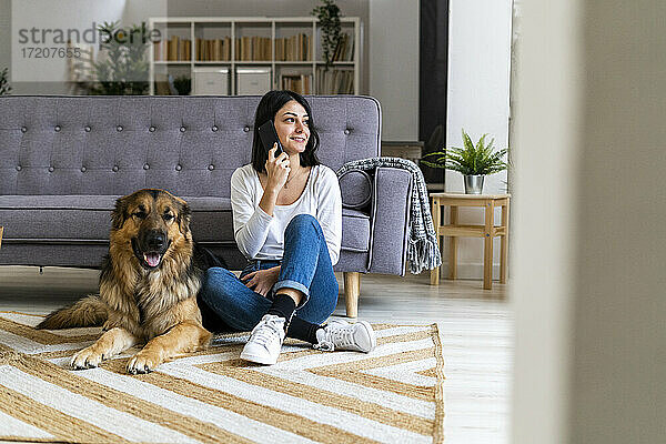 Lächelnde Frau spricht am Telefon  während sie mit ihrem Hund auf dem Teppich zu Hause sitzt