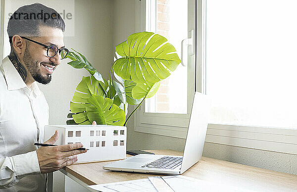 Lächelnder männlicher Architekt  der ein Modell während eines Videogesprächs über einen Laptop am Schreibtisch im Büro hält