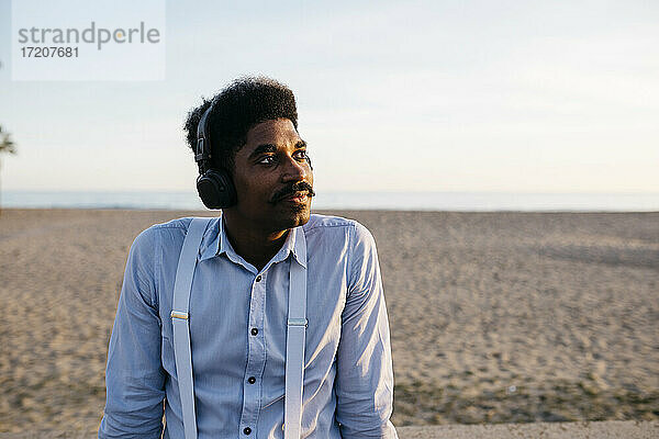 Afro-Mann träumt am Strand und hört Musik über Kopfhörer gegen den klaren Himmel