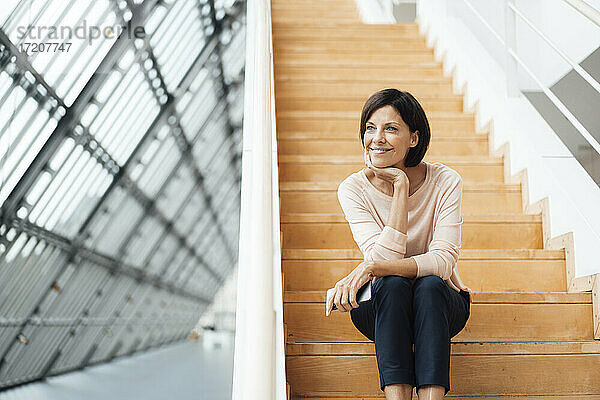 Lächelnde Geschäftsfrau mit Hand am Kinn auf einer Treppe im Korridor sitzend