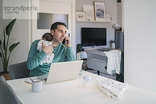 Vater mit Laptop  der seinen Sohn trägt  der wegschaut  während er am Schreibtisch im Heimbüro mit dem Handy telefoniert