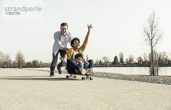 Mann schiebt Frau  die mit einem Jungen auf einem Skateboard sitzt  an einem sonnigen Tag
