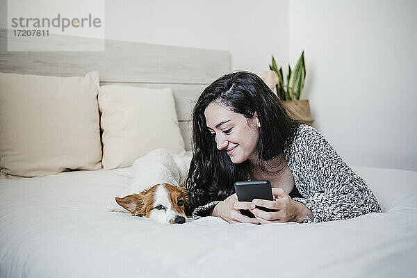 Lächelnde Frau mit Mobiltelefon neben Jack Russell Terrier auf dem Bett liegend