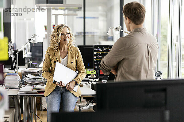 Lächelnde Geschäftsfrau mit Laptop  die mit einem jungen Kollegen diskutiert  während sie sich an einen Schreibtisch im Büro lehnt