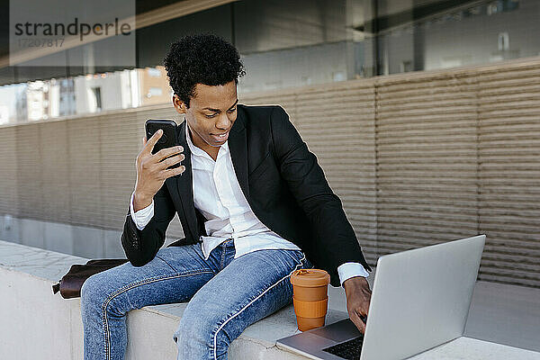 Lächelnder männlicher Unternehmer mit Smartphone und Laptop  der auf einer Stützmauer sitzt