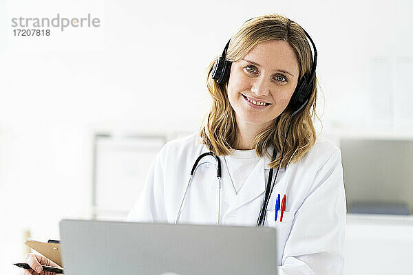 Lächelnde blonde Ärztin mit Kopfhörern während einer Online-Konsultation in einer medizinischen Klinik