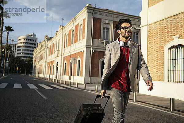 Lächelnder Geschäftsmann  der beim Gehen auf der Straße einen Koffer zieht