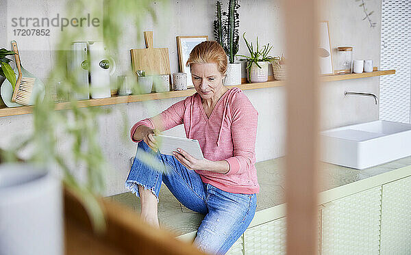 Schöne Frau  die ein digitales Tablet benutzt  während sie auf der Arbeitsplatte zu Hause sitzt