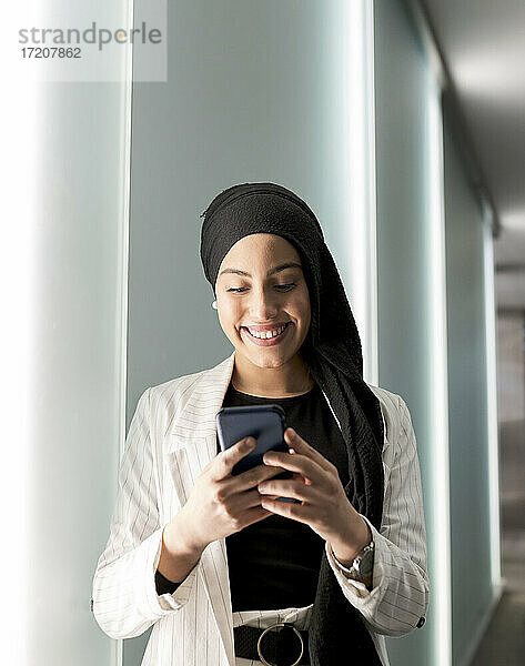 Lächelnde arabische Frau mit Kopftuch und Smartphone an der Wand eines Einkaufszentrums