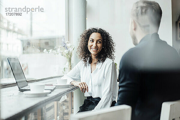 Lächelnde Geschäftsfrau im Gespräch mit einem Kollegen  während sie im Café am Laptop sitzt