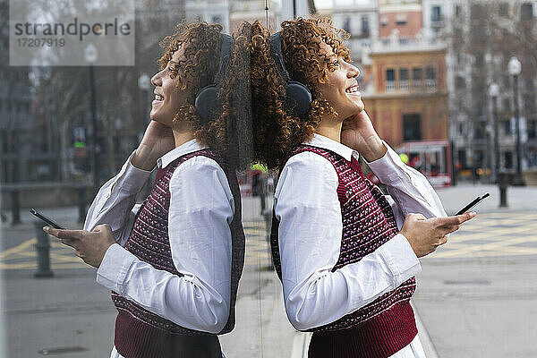 Fröhliche Frau mit Kopfhörern  die mit einem Mobiltelefon an einer Glaswand steht