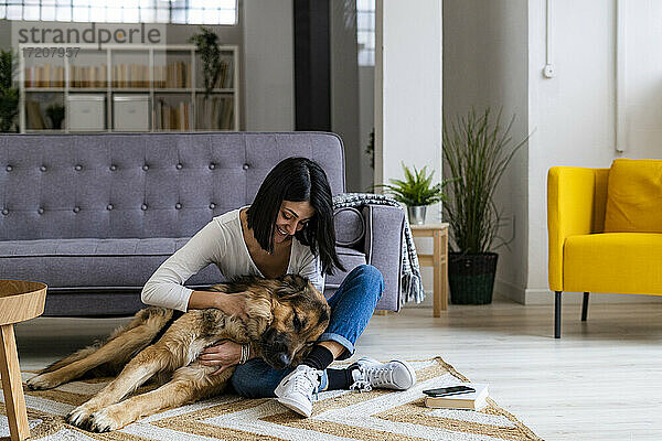 Fröhliche Frau  die ihren Hund umarmt  während sie auf dem Teppich im Wohnzimmer sitzt