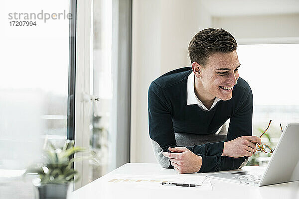 Lächelnder männlicher Berufstätiger  der sich auf einen Stuhl stützt und einen Laptop im Heimbüro betrachtet