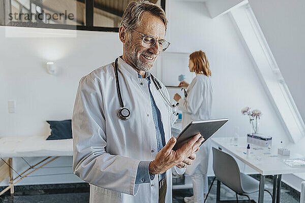Allgemeinmediziner  der ein digitales Tablet benutzt  während er mit einem Mitarbeiter im Hintergrund in einer Klinik steht