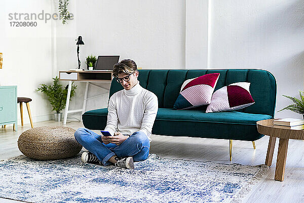 Junger Mann sitzt mit gekreuzten Beinen im Wohnzimmer und benutzt ein Smartphone