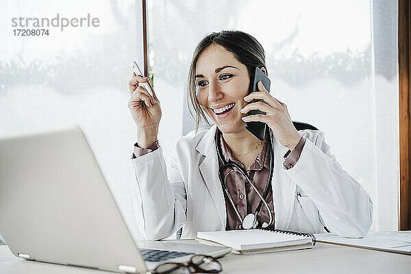 Lächelnde Ärztin  die auf einen Laptop schaut  während sie mit einem Mobiltelefon im Krankenhaus spricht