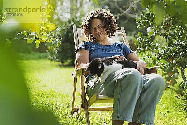 Lächelnde erwachsene Frau  die eine Katze trägt  während sie im Liegestuhl im Garten sitzt