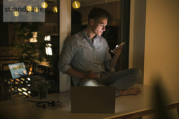 Junger männlicher Unternehmer  der ein Smartphone benutzt  während er zu Hause auf dem Tisch im Wohnzimmer sitzt