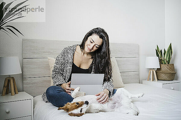 Lächelnde Frau mit Laptop  die einen Jack Russell Terrier streichelt  während sie zu Hause auf dem Bett sitzt