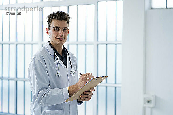 Junger Arzt schreibt Notizen und hält Klemmbrett im Krankenhaus