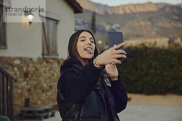 Verspielte Frau streckt die Zunge heraus  während sie ein Selfie mit ihrem Handy vor einem Haus macht