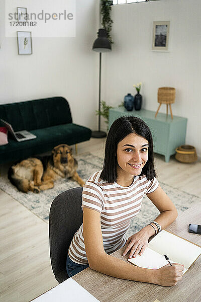 Lächelnde junge Frau mit Stift und Notizbuch an einem Tisch im Wohnzimmer sitzend