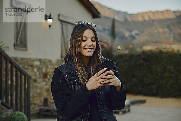 Lächelnde schöne Frau  die ein Mobiltelefon am Haus benutzt