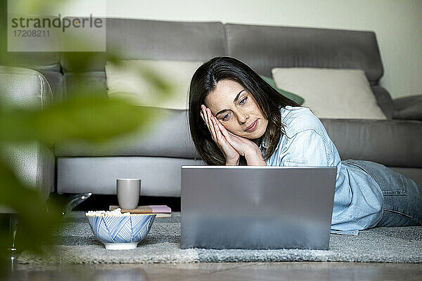 Ernste Frau schaut auf den Laptop  während sie zu Hause auf dem Teppich liegt