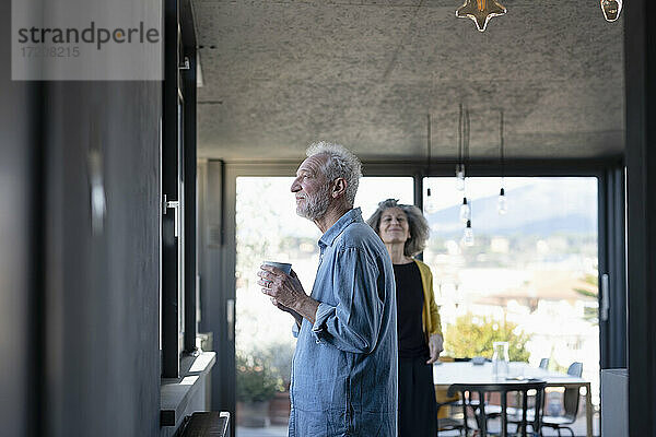 Mann hält Kaffeetasse  während er am Fenster steht  mit Frau im Hintergrund zu Hause