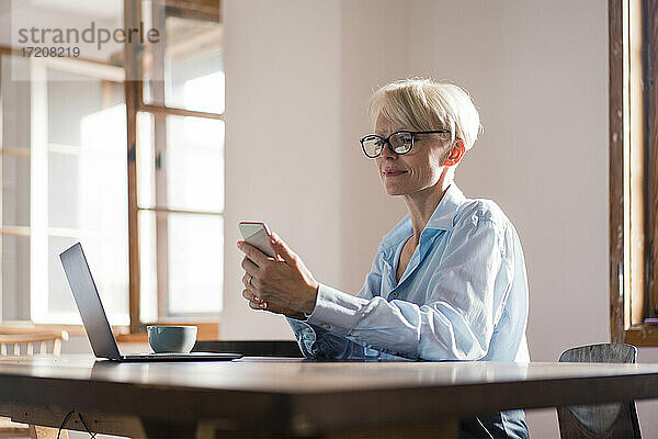 Lächelnde Unternehmerin mit Laptop  die ein Mobiltelefon benutzt  während sie am Schreibtisch im Büro zu Hause sitzt