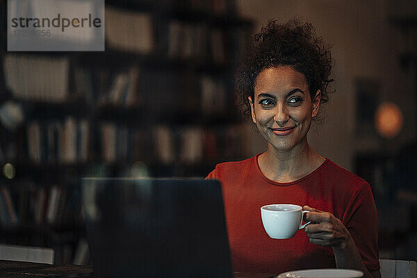 Lächelnde Frau mit Laptop  die eine Kaffeetasse hält  während sie im Cafe sitzt