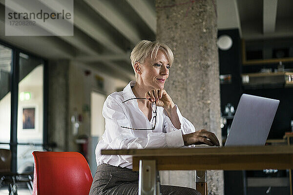 Lächelnde Geschäftsfrau  die eine Brille hält  während sie einen Laptop am Schreibtisch neben einer Säule im Büro zu Hause benutzt