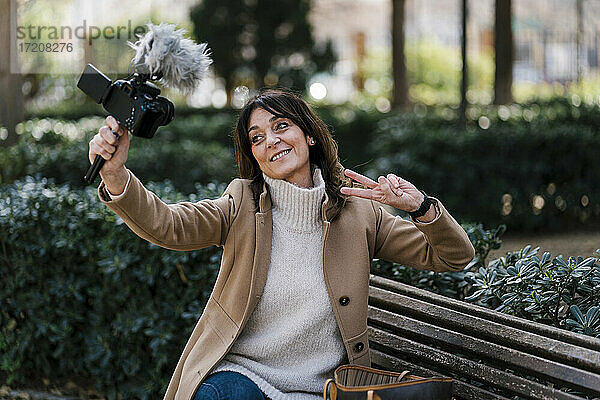 Lächelnde Frau gestikuliert Friedenszeichen  während sie ein Selfie durch die Kamera im Park macht