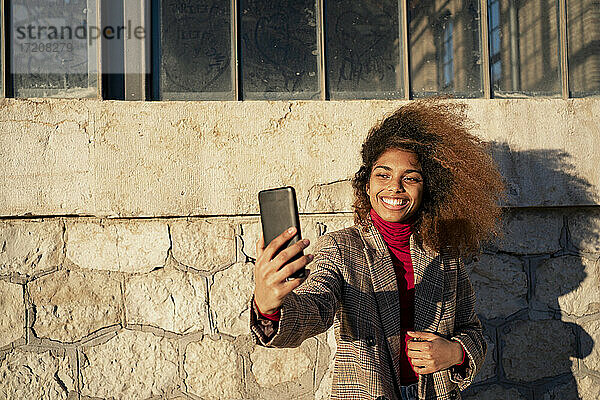 Lächelnde junge Frau  die ein Selfie mit ihrem Smartphone macht  während sie an einem Gebäude steht