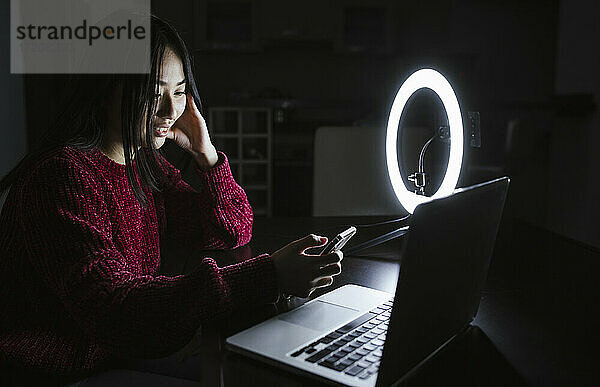 Junge Influencerin  die ihr Smartphone benutzt  während sie mit ihrem Laptop vor dem Ringlicht in der Dunkelkammer sitzt