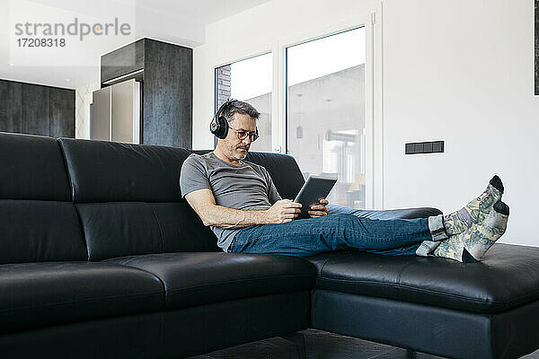 Älterer Mann hört Musik über Kopfhörer  während er ein digitales Tablet im Wohnzimmer benutzt