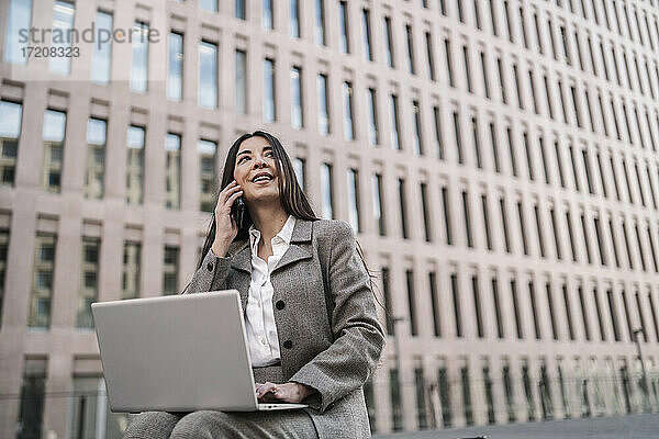 Nachdenkliche Geschäftsfrau  die ein Smartphone benutzt  während sie einen Laptop vor einem Gebäude in der Stadt benutzt