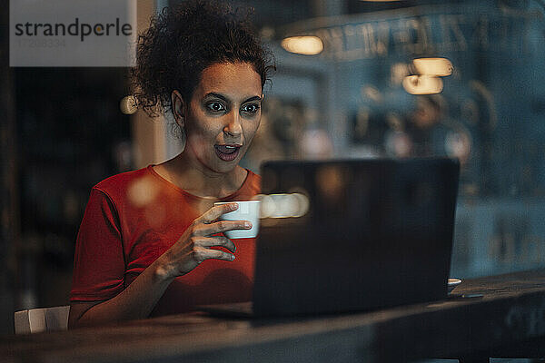Junge Frau hält eine Kaffeetasse und starrt mit offenem Mund auf einen Laptop in einem Café