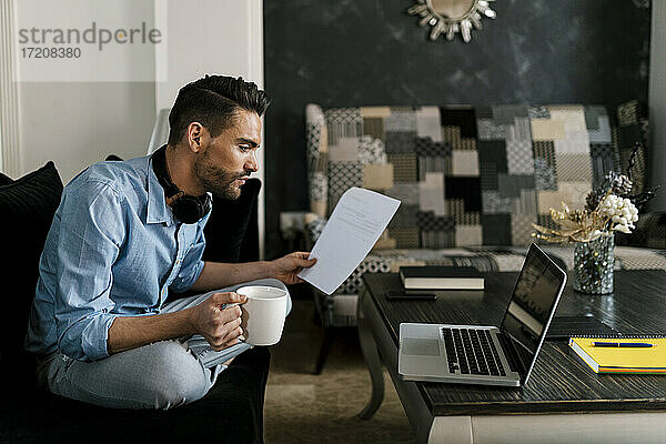 Geschäftsmann mit Kaffeetasse und Papier am Laptop  während er zu Hause sitzt