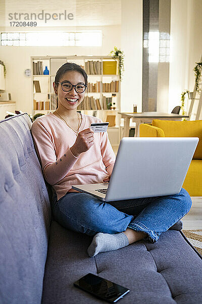 Fröhliche Frau  die eine Kreditkarte in der Hand hält  während sie zu Hause am Laptop sitzt