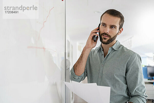 Geschäftsmann  der mit einem Mobiltelefon spricht  während er im Büro vor einem Whiteboard steht