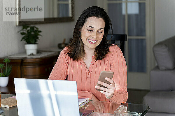 Lächelnde Geschäftsfrau  die auf ihr Handy schaut  während sie am Esstisch im Heimbüro sitzt