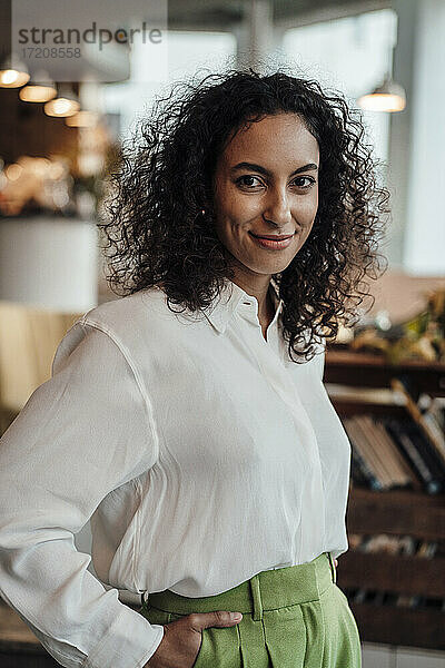 Junge Unternehmerin lächelt  während sie mit den Händen in den Taschen in einem Cafe steht