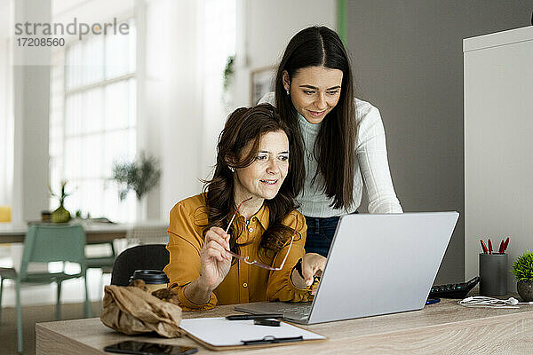 Lächelnde Tochter mit Mutter  die auf einen Laptop schaut  während sie an einem Schreibtisch im Büro zu Hause sitzen