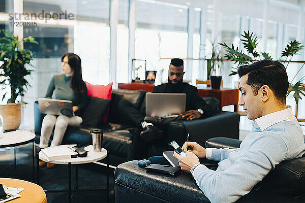 Männlicher Unternehmer bei der Nutzung eines Smartphones während der Arbeit mit Kollegen im Büro