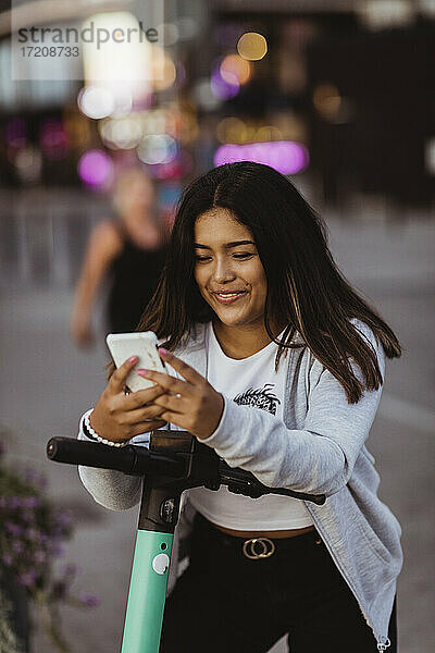 Lächelnde Teenager-Mädchen mit Smartphone im Stehen mit elektrischen Push-Roller